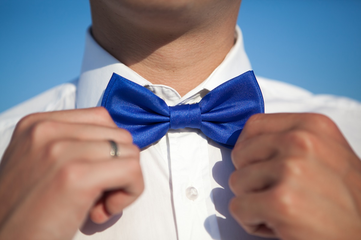 Groom in a white shirt straightens a blue bow tie against a blue sky
