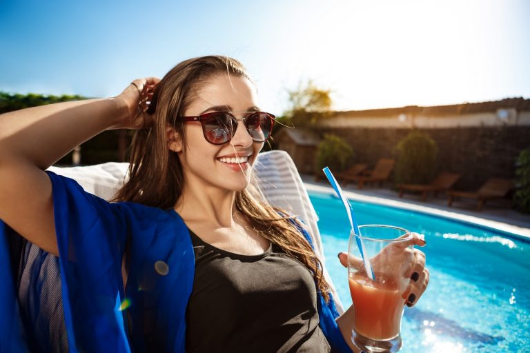 Beautiful girl drinking cocktail, lying on chaise near swimming pool.