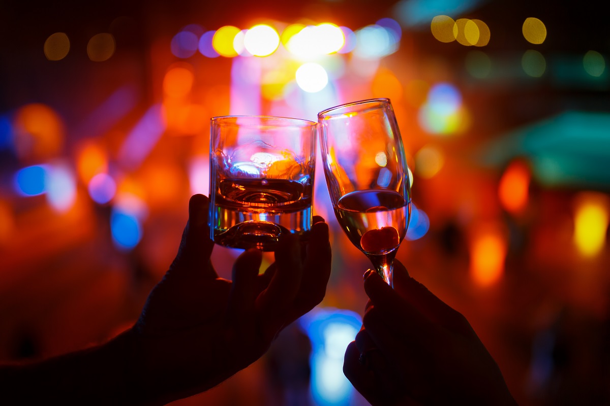 Wineglass of champagne in woman hand and a glass of whiskey in a man hand against a background of colored lights