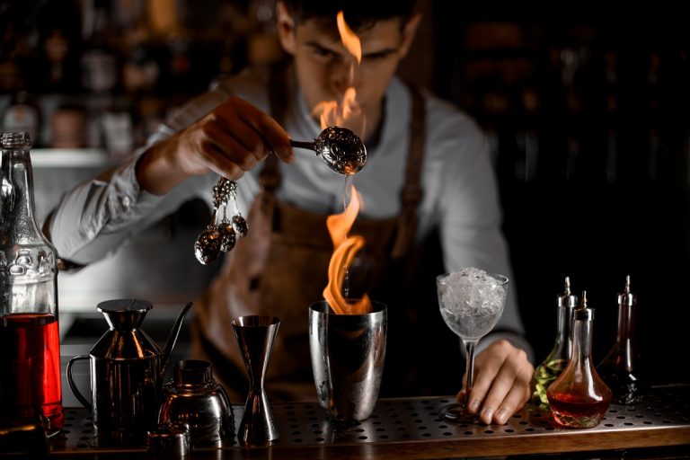 Attractive male bartender pouring an essence from the spoon in the flame to the steel shaker